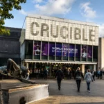 Crucible World Snooker Championship banner above the Crucible Theatre entrance in Sheffield, with fans queuing outside and the snooker player statue in Tudor Square