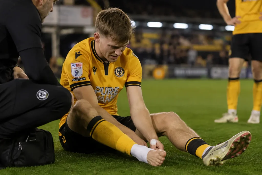 James Crole ankle injury sees Newport striker sit on the pitch clutching his bare foot as a member of staff attends to him during a match at Rodney Parade
